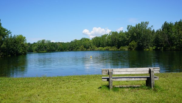 Banc en bois rustique invitant à la détente au bord d'un lac paisible, entouré d'une verdure sous un ciel bleu éclatant à la Base de Plein-Air de Sainte-Foy, Québec.
