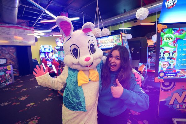 Une personne souriante pose avec un personnage déguisé en lapin de Pâques à l'intérieur de l'arcade Carie Factory, entourée de jeux d'arcade colorés et lumineux