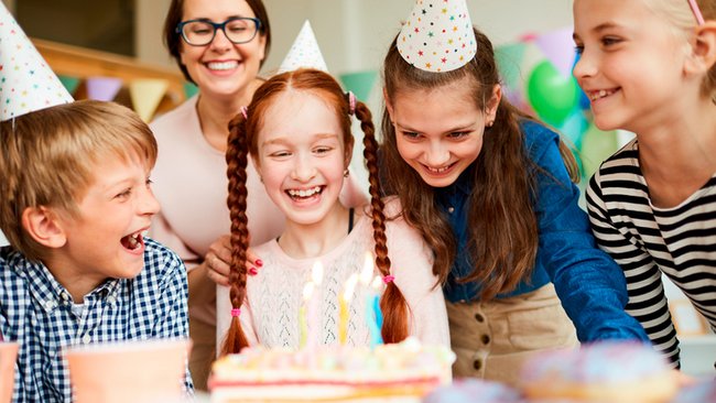 A group of children wearing party hats are smiling and laughing around a birthday cake with lit candles. A woman in the background, also smiling, shares this joyful moment. A festive and colourful atmosphere, ideal for celebrating a birthday at L'Hôtel Québec with the special package.